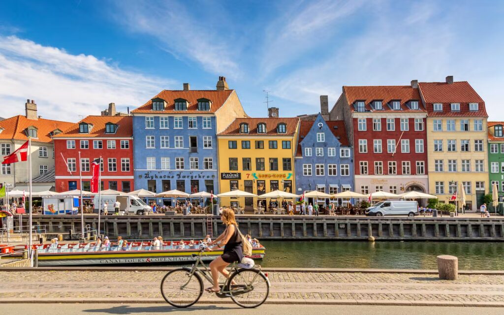 Travel cycling along Copenhagen’s waterfront with a cyclist riding past colorful Nyhavn harbor buildings on a sunny day