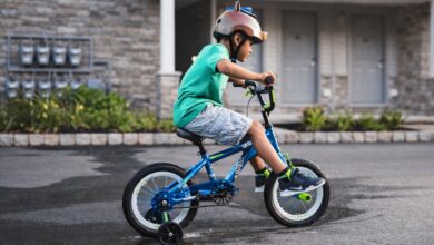 Best Bikes for 2 Year Olds: toddler learning to ride a small blue children’s bike with training wheels while wearing a helmet on a quiet neighborhood street